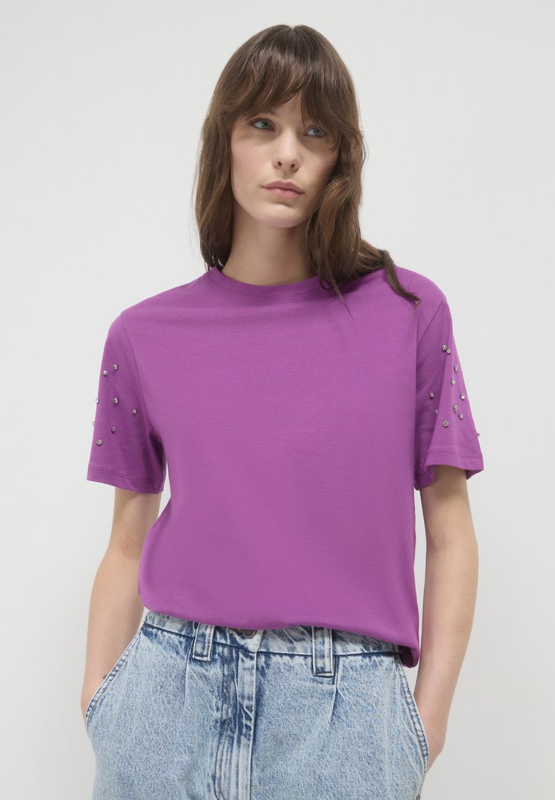 T-shirt viola a maniche corte con collo tondo, caratterizzato da borchie decorative argentate sulle spalle. Realizzato in un tessuto morbido e leggero.
