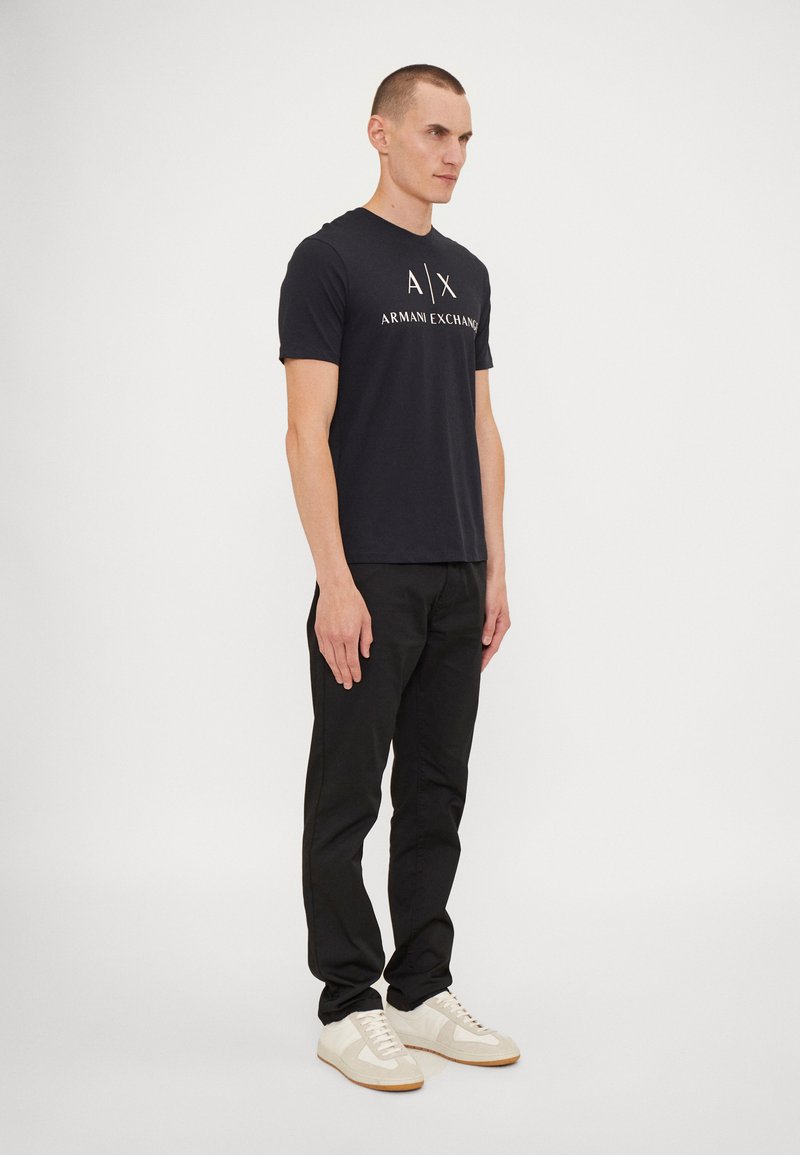 Juodas medvilnės marškinėlis su "ARMANI EXCHANGE" logotipu, derinamas su liesomis juodomis kelnėmis ir šviesiais sportbačiais, modelis stovi prieš baltą foną.