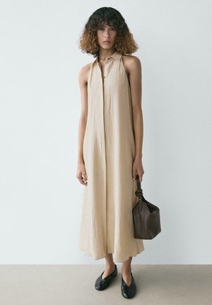 Kobieta o kręconych włosach, ubrana w bezrękawnikową, beżową sukienkę midi i czarne baleriny, trzymająca przy boku ciemnobrązową skórzaną torebkę.