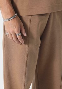 Ruskeat puuvillajoggerit, joissa on teksturoitu pinta ja näkyvät saumat. Hopeaketju ja rengasrannekoru koristavat kättä.