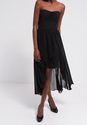 Zwarte strapless jurk met een aansluitend lijfje en een vloeiende high-low zoom. Gemaakt van lichte stof met gerimpelde lagen. Gecombineerd met zwarte hakken.