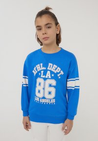 Giovane con capelli legati indossando una felpa blu con strisce bianche e il testo "ATHL. DEPT. LA 86 Los Angeles California", in piedi.