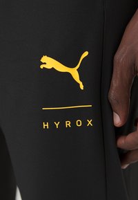 Pantalones deportivos negros con un logo amarillo de Puma y el texto "HYROX". El material es suave y ceñido, diseñado para el rendimiento.