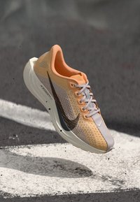 Pantof de alergare Nike portocalie și gri cu swoosh negru, șireturi albe și talpă groasă albă, suspendat deasupra asfaltului cu marcaje albe pe pistă.