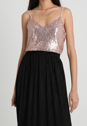 Femme portant un top à bretelles spaghetti rose pailleté associé à une jupe plissée noire taille haute, debout devant un fond uni.