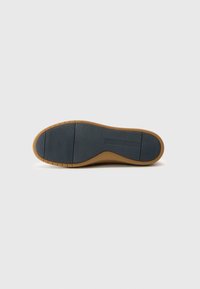 Semelle de chaussure en caoutchouc brun avec un extérieur noir texturé, présentant des rainures horizontales et une surface lisse près du talon, offrant adhérence et durabilité.