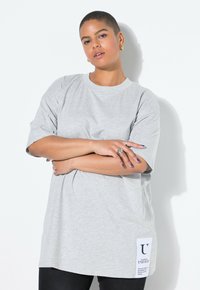 Camiseta gris de gran tamaño hecha de algodón, con mangas cortas y una etiqueta lateral con texto negro. El modelo posa con los brazos cruzados, vistiendo jeans oscuros.