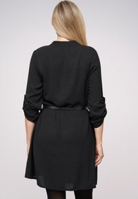 Vrouw met lang blond haar, gekleed in een zwarte jurk met lange mouwen en opgerolde mouwen, met een smalle riem, gezien vanaf de achterkant.