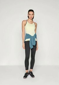 Naistekle, kellel on seljas heledat kollast topi, mustad retuusid, mustad spordijalatsid ja sinine jakk, mis on seotud tema vöökohtade ümber, seismas ühetoonilise tausta ees.