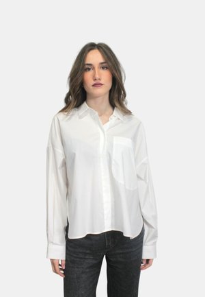 Camicia bianca a maniche lunghe con bottoni in tessuto leggero, dotata di taschino sul petto, orlo arrotondato e vestibilità rilassata. Indossata con jeans scuri.