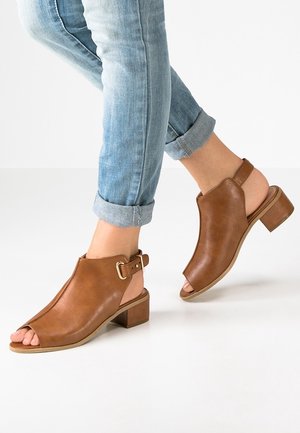 Open-toe brown leather sandals with a chunky heel, featuring a strap and buckle detail. Worn with rolled-up light blue jeans.
