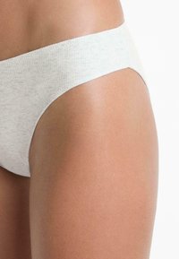 Egy személy csípőjének és felső combjának közeli képe, amely fehér bordás, varrás nélküli alsóneműt visel egy fehér háttér előtt.