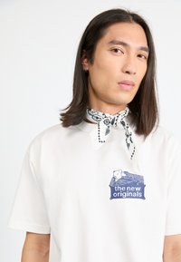 Hvit bomull t-skjorte med et blått grafisk trykk med teksten "de nye originalene." Et svart og hvitt mønstret bandana er bundet rundt halsen.