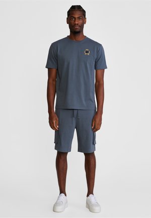 Mann steht da und trägt ein blaues kurzärmeliges Hemd mit einem kleinen Emblem, passende Cargo-Shorts und weiße Turnschuhe vor einem schlichten Hintergrund.