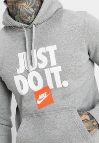 Grå hoodies med framfickor; har vit text "JUST DO IT." och en orange Nike-tag. Mjuk tyg och dragsko i huvan.