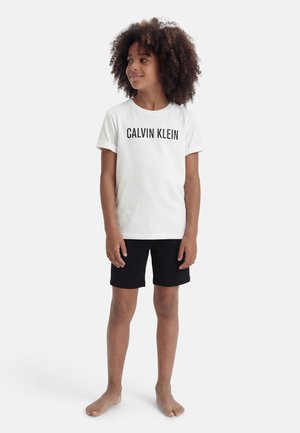 Enfant aux cheveux bouclés portant un t-shirt blanc Calvin Klein et un short noir, debout pieds nus et regardant sur le côté.