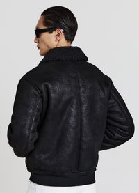 Schwarze Bomberjacke mit strukturierter Oberfläche, ausgestattet mit einem flauschigen Kragen und gerippten Bündchen, die eine lässige, übergroße Silhouette präsentiert.