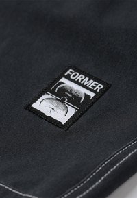 Material textil negru cu o etichetă pătrată cusută care conține cuvântul "FORMER" și grafice abstracte în alb. Cusături contrastante de-a lungul marginilor.