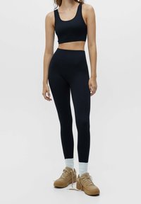 Czarny sportowy crop top i legginsy z wysokim stanem, oba uszyte z gładkiego materiału. Modelka ma na sobie beżowe sneakersy z jasnoniebieskimi skarpetkami.