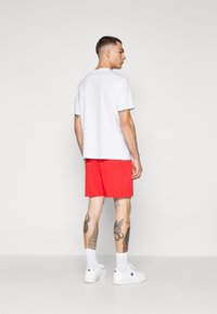 Λευκή μπλούζα, κόκκινες βερμούδες και λευκά αθλητικά παπούτσια. Ορατά τατουάζ στα πόδια. Απλός, σπορ σχεδιασμός σε ζωντανά χρώματα.