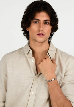 Camicia button-up in lino beige chiaro con taschino sul petto, indossata con un bracciale argentato con sezioni testurizzate e lisce.