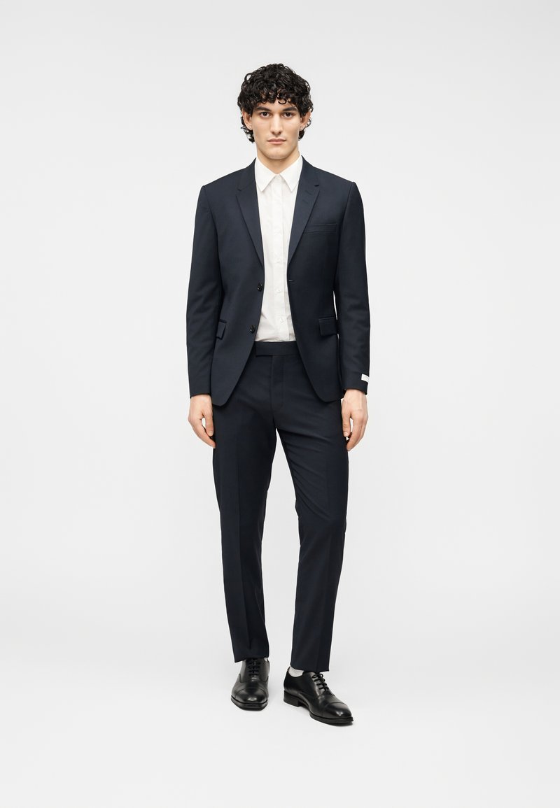 Σκούρο μπλε κοστούμι με σακάκι μον όπετο και παντελόνια στενής γραμμής, συνδυασμένο με λευκό πουκάμισο και μαύρα δερμάτινα παπούτσια. Απλός, ραμμένος σχεδιασμός.