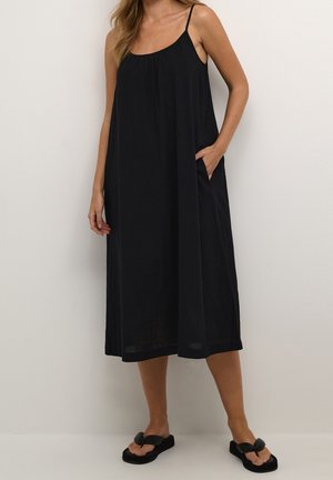 Mujer con un vestido midi negro y suelto de tirantes finos, de pie con una mano en el bolsillo, combinado con sandalias negras de plataforma.