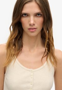 Beige ribbet tank top med en scoop halsudskæring og seks knapper ned foran. Materialet ser blødt ud med en let tekstur.