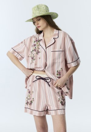 Vrouw draagt een pastelroze gestreept pyjamaset met bloemrijke borduursels en een lichtgroene brede hoed, terwijl ze de zoom van haar hemd rechtzet.
