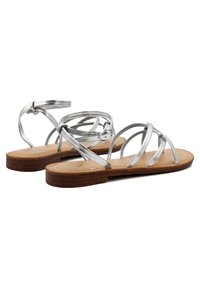 Sandali metallici argento con cinghie incrociate, cinturino alla caviglia con fibbia e suola piatta marrone. Texture liscia e design minimalista.