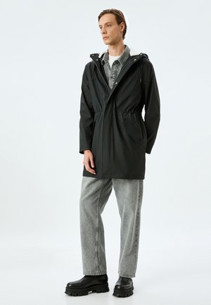 Parka nera impermeabile con vita regolabile, cappuccio e tasche laterali. Indossata sopra una camicia grigia chiara con bottoni e pantaloni in denim grigi.