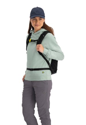 Zwarte rugzak met verstelbare schouderbanden en gele hardware, gedragen over een lichtgroene hoodie en grijze broek. Model heeft lang haar en een marineblauwe pet.