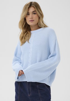 Žena s dlhými blond vlasmi, oblečená v svetlomodrom pletenom svetri s širokými rukávmi a tmavomodrých nohaviciach, stojaca pred jednoduchým pozadím.