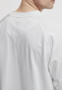 Weißes Langarmshirt aus weichem Stoff, mit rundem Ausschnitt und geprägtem Logo-Detail am oberen Rücken in der Nähe des Ausschnitts.