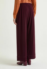 Pantaloni a gamba larga in tessuto bordeaux intenso con una texture liscia. Presentano una vita elasticizzata e polsini ripiegati alla caviglia.