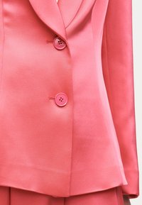 Ροδακινί ροζ σακάκι με έντονη ραφή, δύο στρογγυλά κουμπιά και γιακά με σχισμές, εμφανίζοντας τη μέση και μέρος των ταιριαστών παντελονιών.