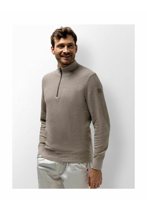 Man wearing light brown quarter-zip sweater and white pants, smiling and looking to the side against a plain white background.