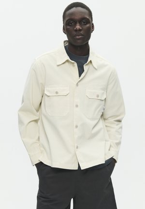 Young person with short hair wearing a cream button-up shirt over a dark top, standing with hands in black pants pockets against a plain background.