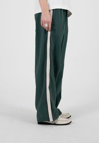 Person iført mørkegrønne bukser med vide ben og hvide striber på siden, hvid top, hvide mokkasinstil sko og en sølvring på venstre hånd.