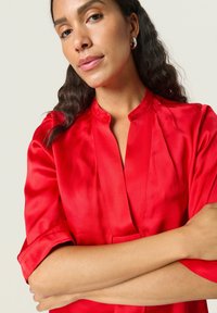 Blusa de satén rojo con cuello mao y mangas cortas. Textura suave y ligero brillo, con un sutil detalle de pliegue en la parte delantera.
