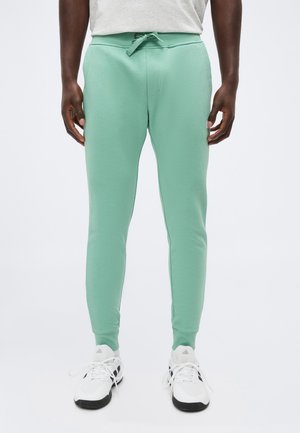 Άντρας που φοράει ανοιχτό πράσινο στενό παντελόνι τζόγκινγκ, λευκά αθλητικά παπούτσια με μαύρες σόλες και λευκό πουκάμισο, όρθιος μπροστά σε απλό φόντο.