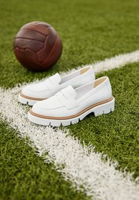 Białe skórzane lordsy z masywną, żłobioną podeszwą i brązowym akcentem, umieszczone na sztucznej trawie obok brązowej piłki nożnej.