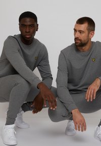 Des sweatshirts et pantalons gris en tissu doux, avec des accents de logo jaunes, sur deux hommes accroupis devant un fond uni.