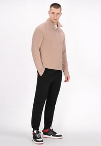 Beige zip-pullover med krave og lange ærmer, parret med sorte tætsiddende bukser og sort/røde sneakers med hvide såler. Glat tekstur.