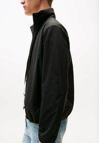 Zwarte lichtgewicht jas met een hoge kraag, ritsluiting en elastische manchetten. Voorzien van een tonale "TOMMY JEANS"-logo langs de mouw.