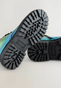 Semelle de chaussure en caoutchouc noir avec un motif de crampon profond ; s'adapte parfaitement contre un matériau supérieur irisé bleu-vert présentant une texture lisse.