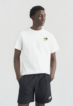 Jonge man die een wit t-shirt van The North Face en zwarte korte broek draagt, staand met handen in de zakken, opzij kijkend tegen een effen achtergrond.