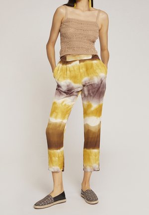 Gehaakte tanktop in lichtbruin; tie-dye broek in geel, bruin en paars; zwarte instap schoenen met een geometrisch patroon.