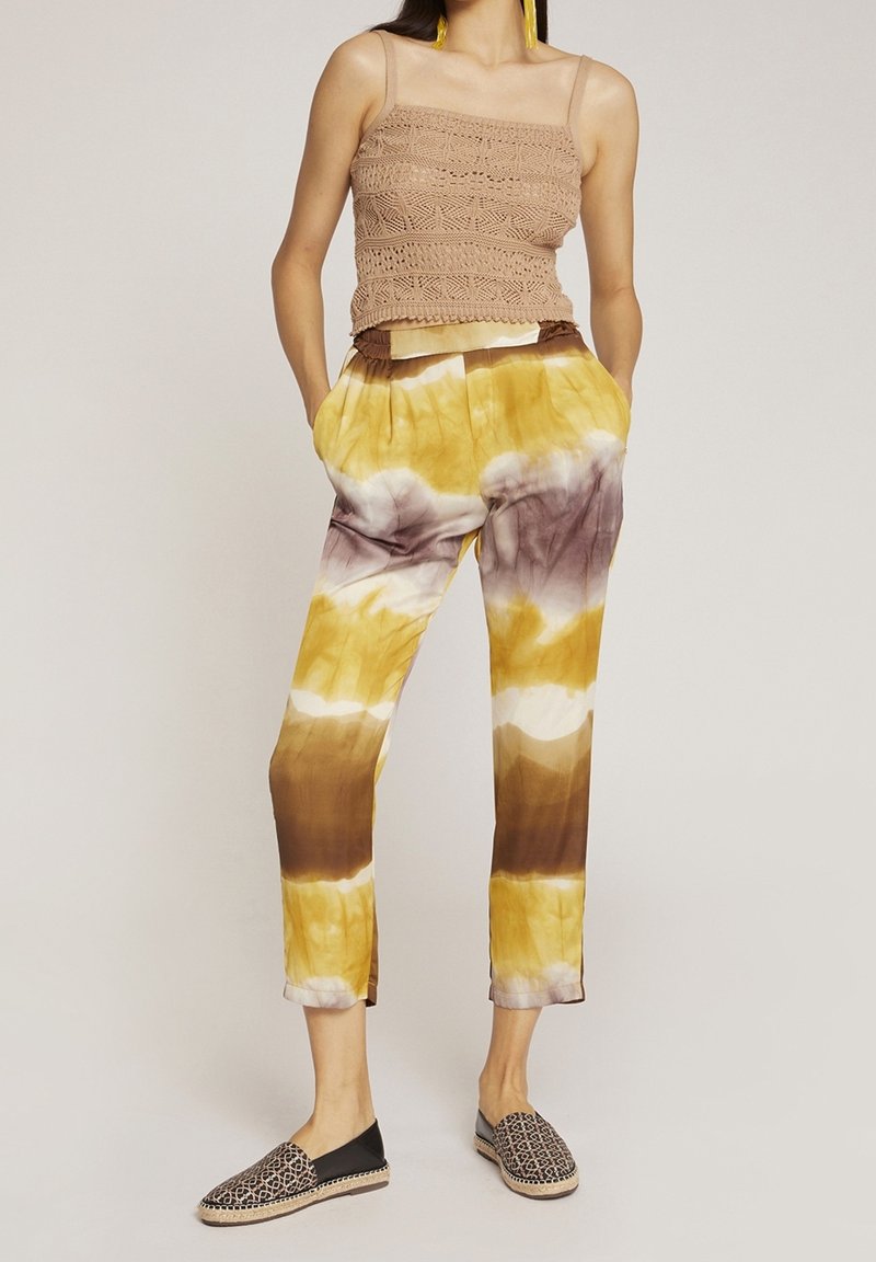 Häklett linne i ljusbrun; batikbyxor i gult, brunt och lila; svarta slip-on skor med ett geometriskt mönster.