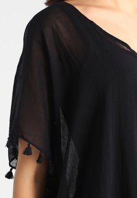 Black, lightweight top featuring a V-neckline, loose fit, and short sleeves. Accented with tassels at the sleeve edges and textured fabric.